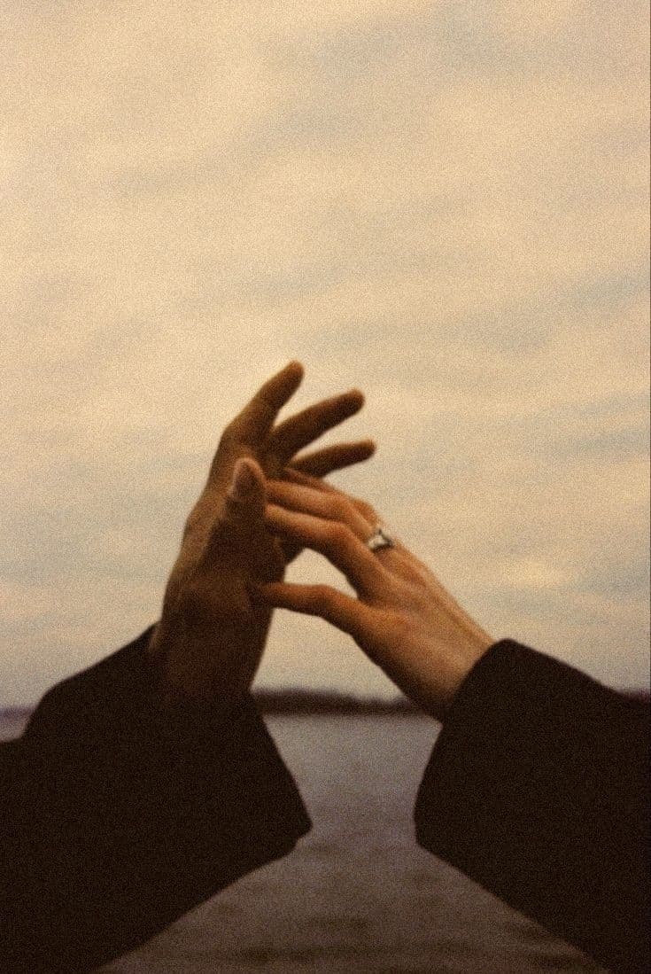 Two hands reaching for each other against a pale, overcast sky above the water, one of them wearing a ring